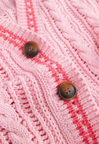 Nahaufnahme eines rosa gestrickten Stoffs mit Zopf- und Rippenmustern, der zwei braune runde Knöpfe auf einem rot gestreiften Abschnitt aufweist.