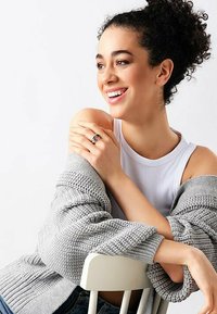 Cardigan grigio a maglia indossato sopra una canotta bianca, seduto su una sedia, con un anello d'argento su una mano e capelli ricci.