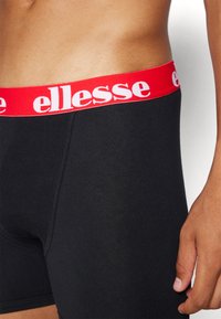 Svarta boxertrosor med ett rött elastiskt midjeband som har den vita "ellesse"-logotypen. Tyget är mjukt och elastiskt, designat för komfort.