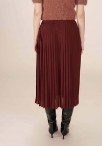 Jupe midi plissée couleur vin avec taille élastique, associée à des bottes noires montantes, vue de dos. Texture de tissu lisse.