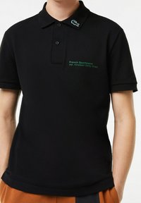 Polo noir en tissu texturé, avec un logo vert brodé sur le col et la poitrine, accompagné d'un texte indiquant l'année de création.