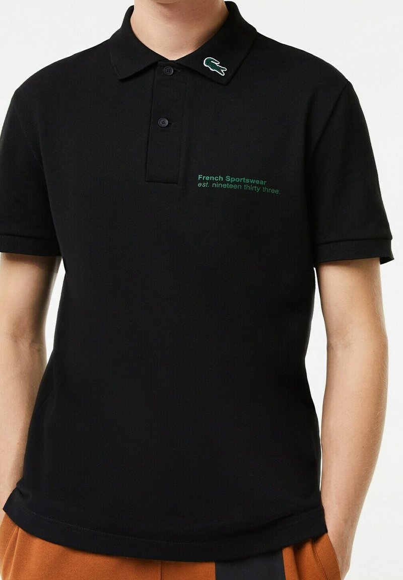Polo noir en tissu texturé, avec un logo vert brodé sur le col et la poitrine, accompagné d'un texte indiquant l'année de création.