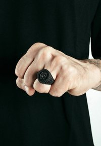 Anillo negro con un diseño en espiral, superficie texturizada, llevado en una mano con un puño cerrado. El fondo es oscuro y uniforme.