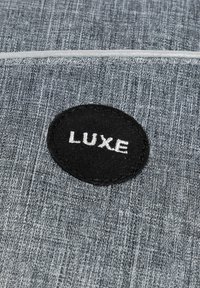 Grauer Stoff mit einer strukturierten Oberfläche zeigt ein rundes schwarzes Patch, auf dem das Wort "LUXE" in Weiß gestickt ist.