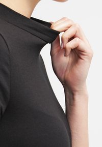 Svart, åtsittande topp med hög krage, mjuk textur och slät finish. En hand lyfter försiktigt kragen för att avslöja innersömmen.