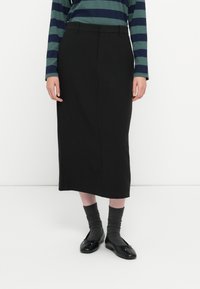 Jupe noire midi avec une texture lisse, coupe droite et détails de couture minimalistes ; associée à de longues chaussettes grises et des ballerines noires.
