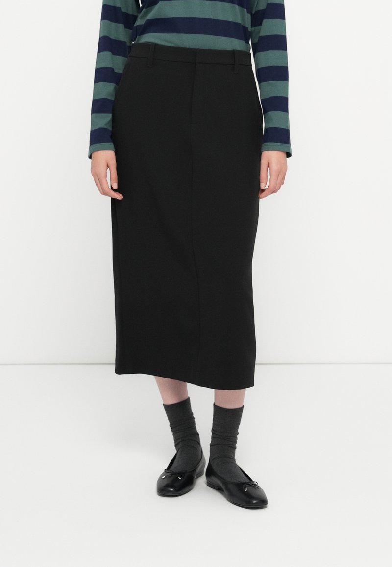 Jupe noire midi avec une texture lisse, coupe droite et détails de couture minimalistes ; associée à de longues chaussettes grises et des ballerines noires.