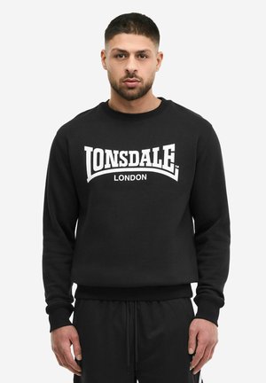 Homme de face portant un sweat-shirt noir avec l'inscription « Lonsdale London » en blanc sur la poitrine.