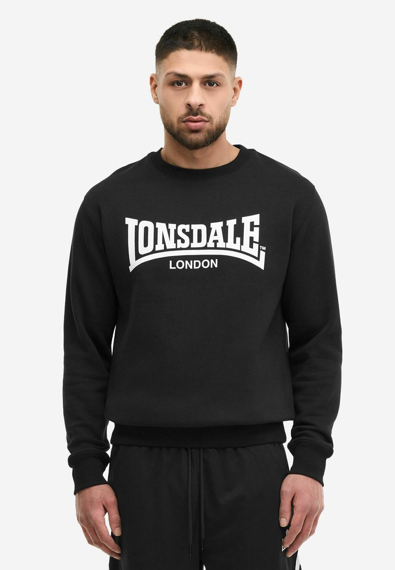 Homme de face portant un sweat-shirt noir avec l'inscription « Lonsdale London » en blanc sur la poitrine.