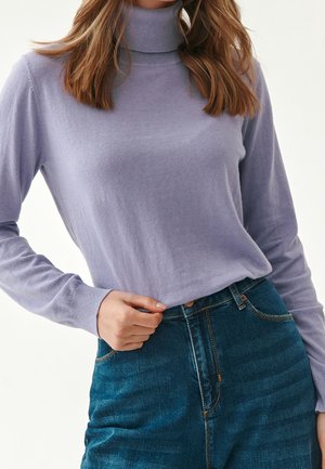 Femme portant un pull à col roulé violet clair associé à un jean en denim bleu foncé taille haute, main posée près de la taille.