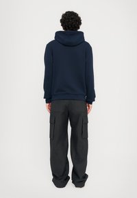 Marineblå hettegenser med stor hette og ribbestrikkede mansjetter, kombinert med løstsittende svarte cargo-bukser med flere lommer. Stoffet ser mykt ut.