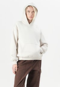 Model with blonde hair wearing a light beige hoodie and brown pants, standing against a plain white background, looking directly at the camera.