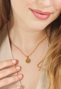 Gouden ketting met een cirkelvormige hanger waarop een gegraveerde "M" staat, gedragen tegen een neutraal gekleurde halslijn.
