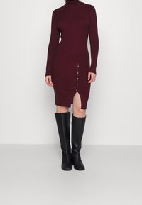 Burgundská pletená šaty s vysokým výstřihem, bočním rozparkem a zlatými knoflíky. V kombinaci s vysokými černými botami pro kontrast.