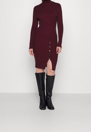 Bordeaux ribbelgebreide jurk met een hoge halslijn, een zijsplit en gouden knopen. Gecombineerd met hoge zwarte laarzen voor contrast.