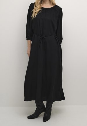 Kvinde iført en lang sort kjole med 3/4-ærmer og bindebælte i taljen, kombineret med sorte støvler med hæl, stående foran en ensfarvet baggrund.