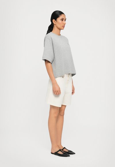 Camiseta corta de manga corta gris, pantalones cortos blancos de cintura alta y zapatos negros tipo slip-on, mostrados en un modelo de pie de perfil.