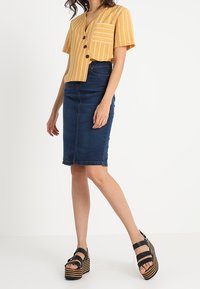 Camiseta de manga corta a rayas amarillas con escote en V y bolsillo, combinada con una falda lápiz de mezclilla y sandalias de plataforma negras con suelas a rayas.