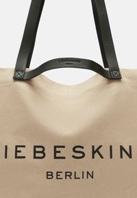 Beige Canvas-Shopper mit schwarzen Ledergriffen und bedrucktem Logo "LIEBESKIND BERLIN." Glatte Textur, minimalistisches Design.