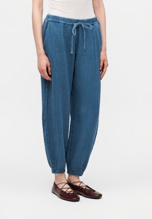 Γυναίκα που φορά μπλε υφασμάτινο παντελόνι με κορδόνι και σκουροκόκκινα επίπεδα δερμάτινα παπούτσια με κορδόνια, στέκεται μπροστά σε απλό ανοιχτό φόντο.