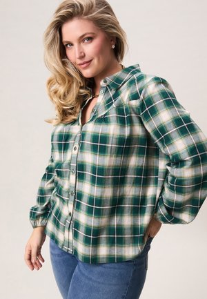 Vrouw met lang blond haar, gekleed in een groen-wit geruit overhemd en een blauwe spijkerbroek, glimlachend en poserend met haar hand op haar heup.