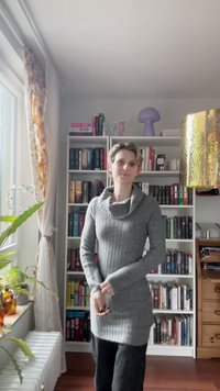Šedý žebrovaný svetr s rolákem, spárovaný s černými kalhotami. V pozadí jsou knihovny, rostlina a dekorativní předměty.