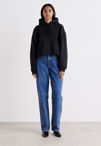 Pullover corto nero con cappuccio e un logo discreto, abbinato a jeans dritti blu a vita alta e scarpe nere con punta.