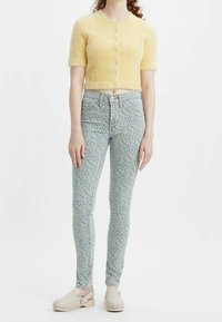 Cárdigan corto de color amarillo y peludo con mangas cortas, combinado con jeans ajustados de patrón floral en azul claro y zuecos beige.