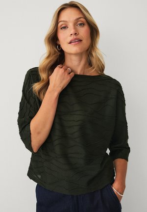 Kobieta o falowanych blond włosach, ubrana w ciemny, teksturowany top z rękawem do łokcia i ciemne spodnie, dotykająca dekoltu lewą ręką.