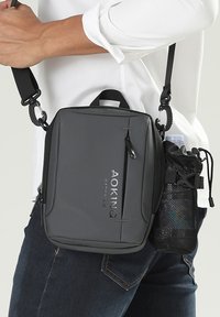Borsa a spalla grigia con design rettangolare, tasca con zip e marchio "AOKING". Portabottiglie in rete attaccato. Cinghia regolabile visibile.