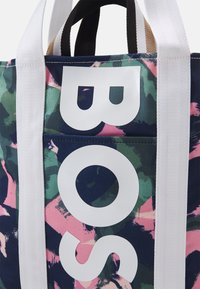 Sac en toile avec un motif abstrait bleu marine, vert et rose. Présente un texte blanc et des poignées blanches robustes. Comprend une poche à patch.