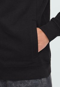 Hånd delvis satt inn i frontlommen på en svart hettegenser, iført mørkegrå jeans.