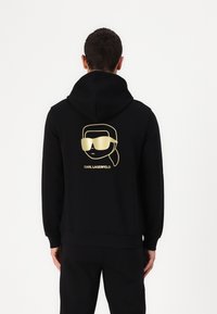 Felpa nera con cappuccio che presenta una grafica dorata di un personaggio stilizzato con occhiali da sole sulla schiena. Il materiale sembra morbido e ha una vestibilità standard.