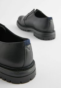 Zapatos de cuero negros con un acento de tela azul texturizada en el talón, que presentan una suela negra gruesa y robusta, y un diseño elegante y minimalista.