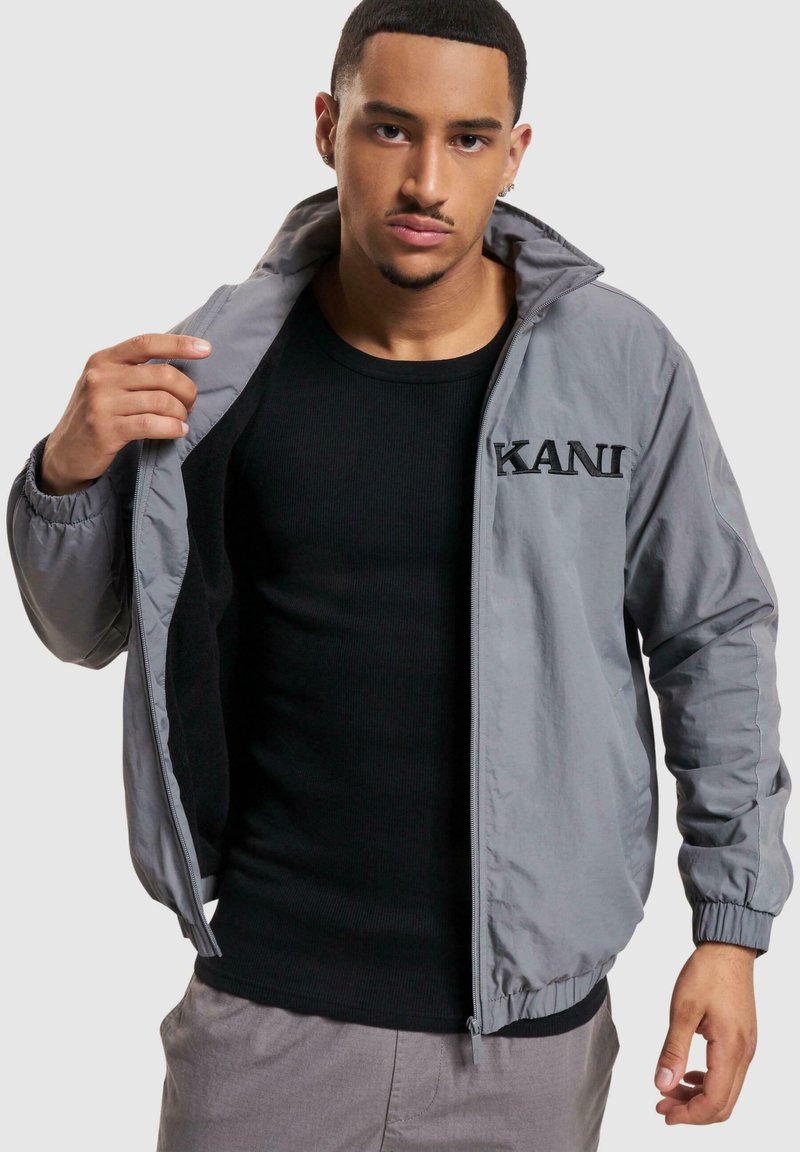 Lightweight gray jacket with a zip front, ribbed cuffs, and embroidered "KANI" logo. Worn over a black fitted shirt. Smooth fabric texture.