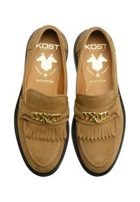 Mocassins en daim marron avec des détails en frange et une accent en chaîne dorée. Semelle en caoutchouc noir, texture lisse et design à bout arrondi.