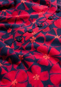 Gėlių raštuotas audinys, kuriame yra dideli raudoni ir oranžiniai žiedai tamsiai mėlyname fone. Šeši juodi mygtukai išdėstyti vertikaliai. Minkšta tekstūra.