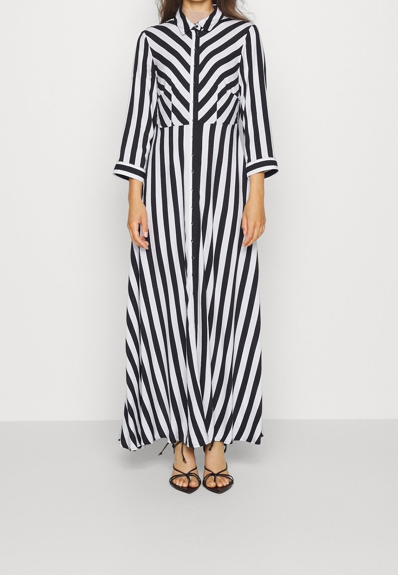 Femme portant une longue robe chemise rayée noire et blanche avec des manches trois-quarts et des sandales noires à brides.