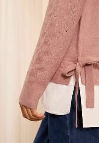 Roza pletena dukserica s teksturiranim rukavima u obliku srca, s remenom u struku i rebrastim manžetama, složena preko bijele košulje.