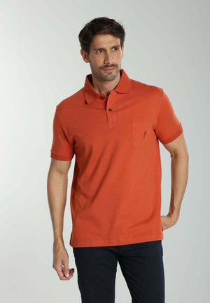 Homme portant un polo orange à manches courtes avec poche et pantalon foncé, debout devant un fond clair uni, regardant légèrement vers la droite.