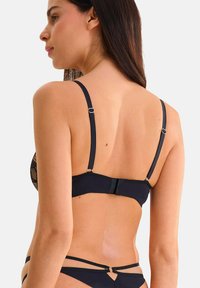 Femme portant un soutien-gorge en dentelle noire et des sous-vêtements assortis avec lanières, vue de dos contre un fond clair uniforme.