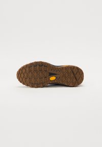 Terrengløpesko såle, med en robust brun gummisåle og vinkelformede mønstre. Merkbar gul Vibram-logo til stede.
