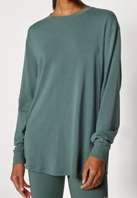 Långärmad tröja med rund halsringning i dämpad grön, tillverkad av mjukt material; har en avslappnad passform och ribbade ärmslut, med sömnadsdetaljer vid nederkanten.