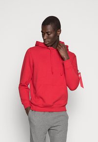Felpa rossa realizzata in tessuto misto cotone, con tasca davanti, cappuccio regolabile con cordino e etichetta laterale. Abbinata a pantaloni grigi.