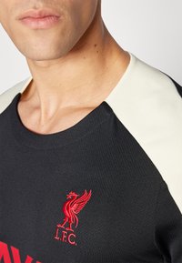 Maillot de sport noir avec le logo rouge de Liverpool FC ; présente des accents crème sur les épaules et un tissu texturé. Design à col rond.