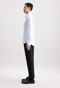 Camicia button-up a righe blu chiaro, infilata in pantaloni navy su misura. Scarpe eleganti nere. Figura mostrata di profilo su uno sfondo neutro.