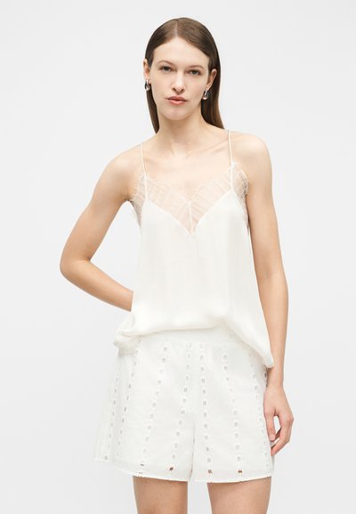 Młoda kobieta w białej, bezrękawnikowej koszulce na ramiączkach z koronkowym wykończeniem oraz białych szortach z ażurowym wzorem, stojąca z ręką na biodrze na jednolitym tle.