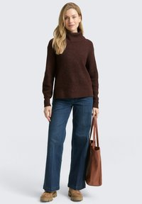 Brauner strukturierter Strickpullover mit Rollkragen und langen Ärmeln, kombiniert mit blauen weiteren Jeans und beigen Plateauschuhen, hält eine braune Tote-Tasche.