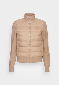 Beige pufferjack met een gebreide trui-lichaam, hoge kraag, rits aan de voorkant en logo patch. Gladde textuur op het puffer-gedeelte, geribde manchetten.