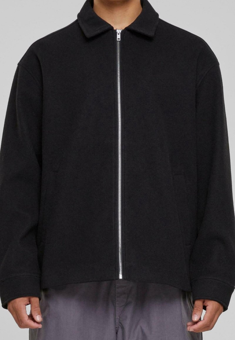 Svart zip-up jacka med krage, tillverkad av mjukt material. Har en rak skärning och sidofickor. Den släta dragkedjan kontrasterar mot tygtexturen.